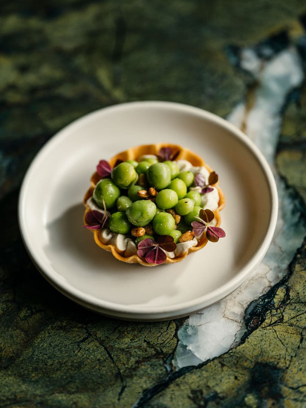 A tart topped with green peas and micro herbs on a white plate with a dark marble background.