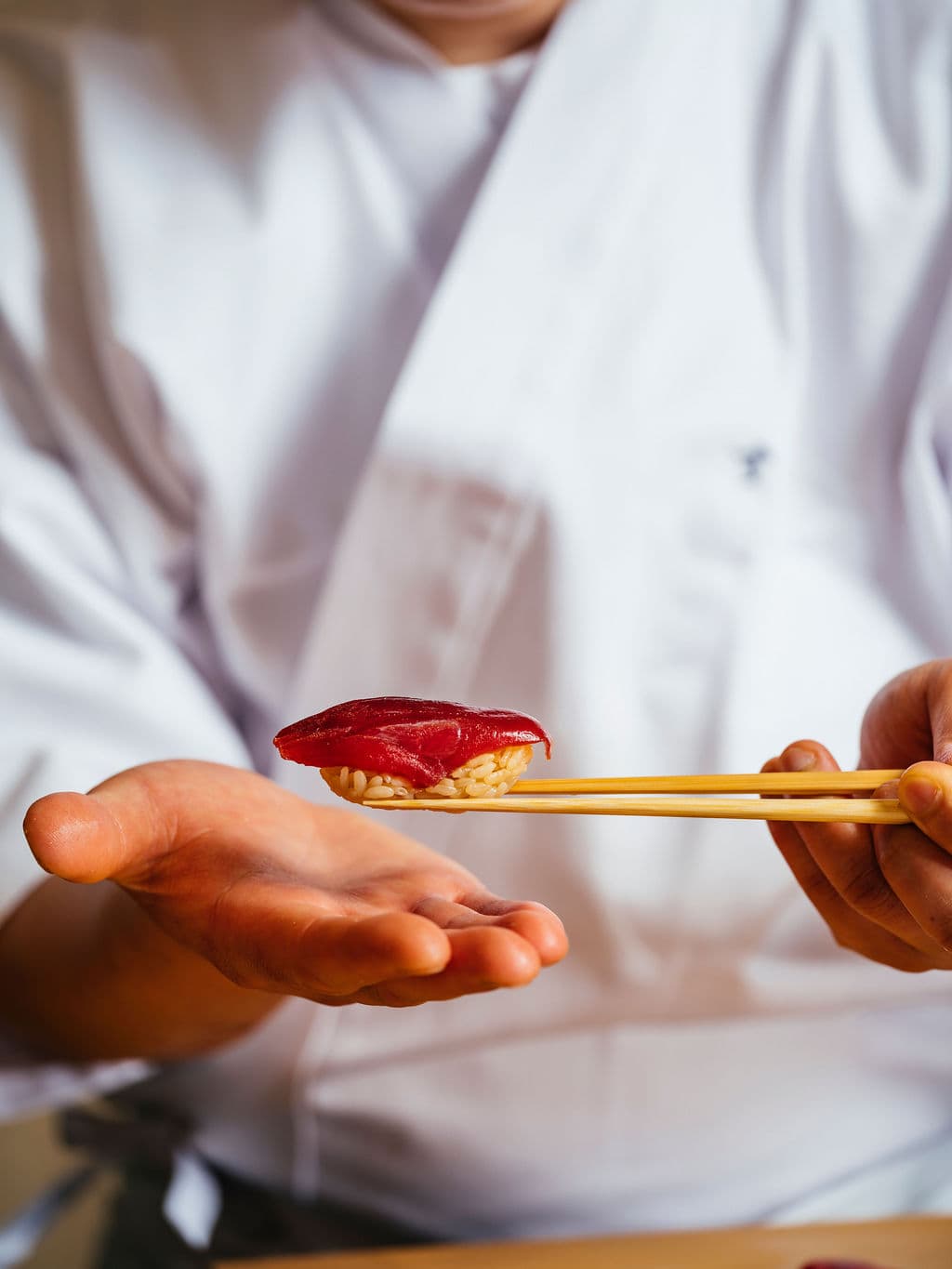A chef presenting a piece of tuna sushi held with chopsticks above his hand.