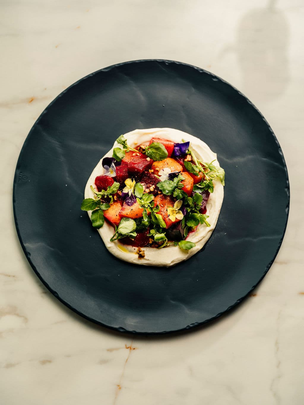 A black plate with a dish of fresh strawberries, herbs, and edible flowers on a marble table.