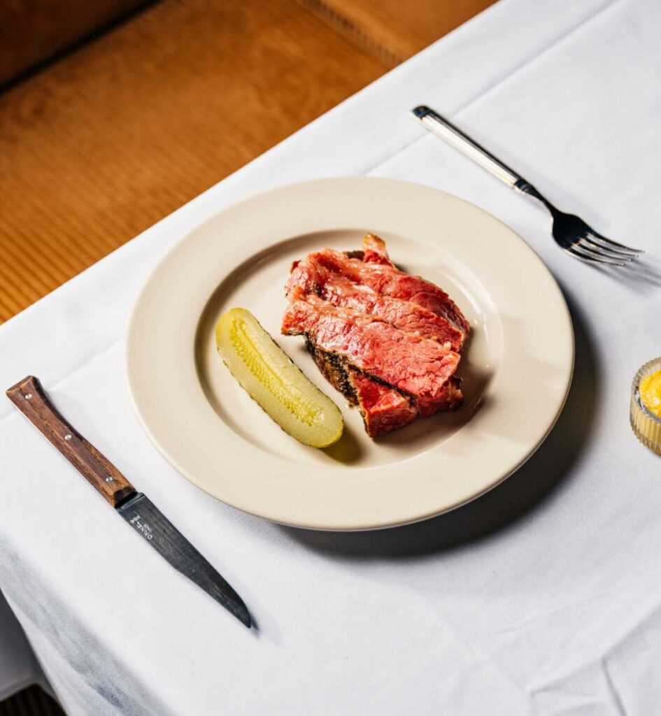 Plate of smoked meat with a pickle spear, served on a white tablecloth.